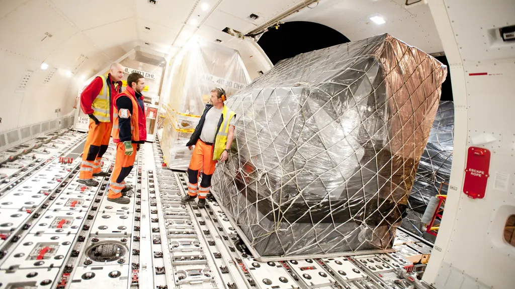 A netted air cargo pallet standing on the roller floor of a freighter aircraft's main deck, with ground handling staff in high-visibility clothing beside it.