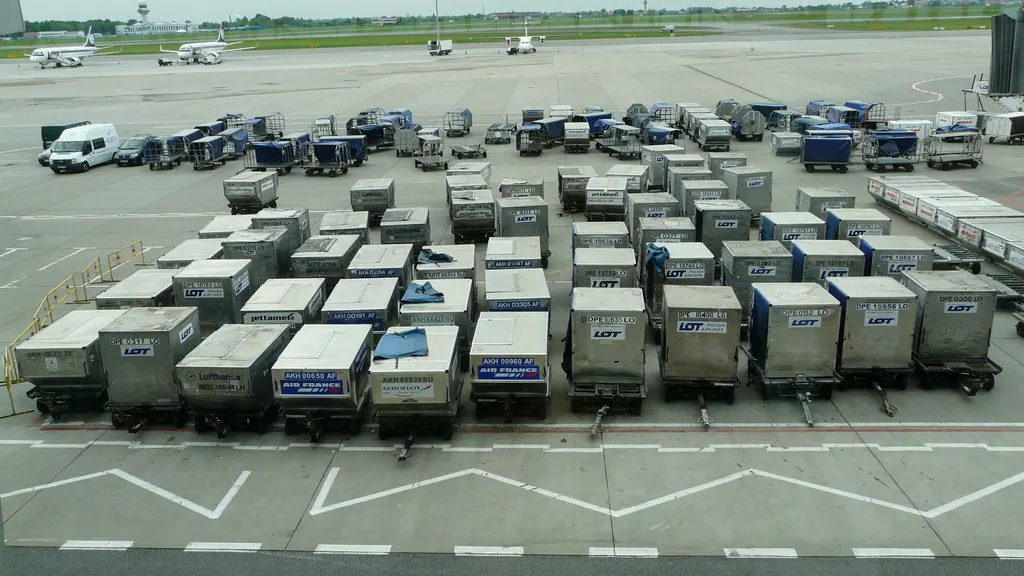 Rows of air cargo containers standing on their dollies on an airport apron, with an airfield and terminal buildings behind them.