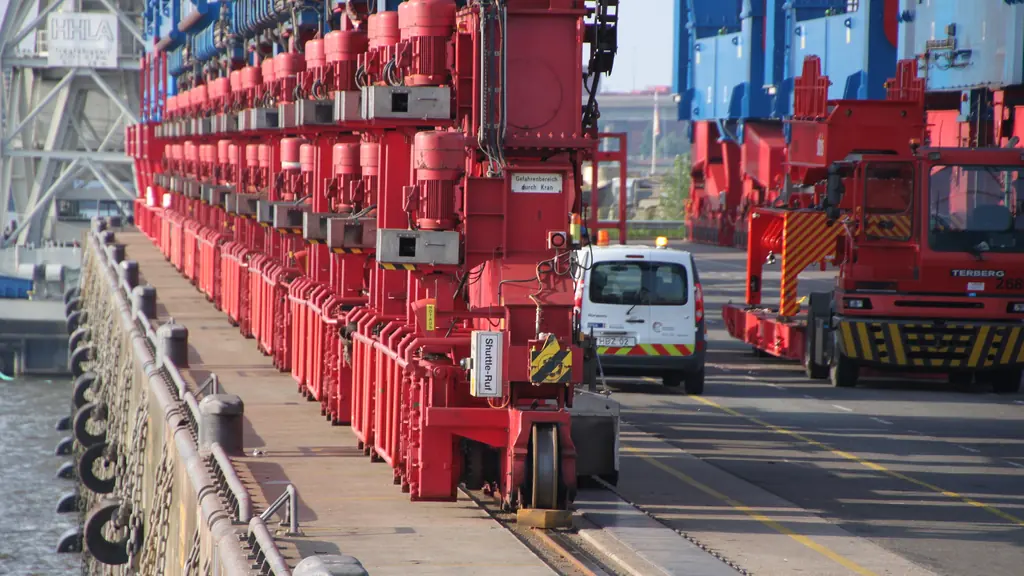 A row of red container gantry cranes along a quay, with terminal tractors working beneath them.