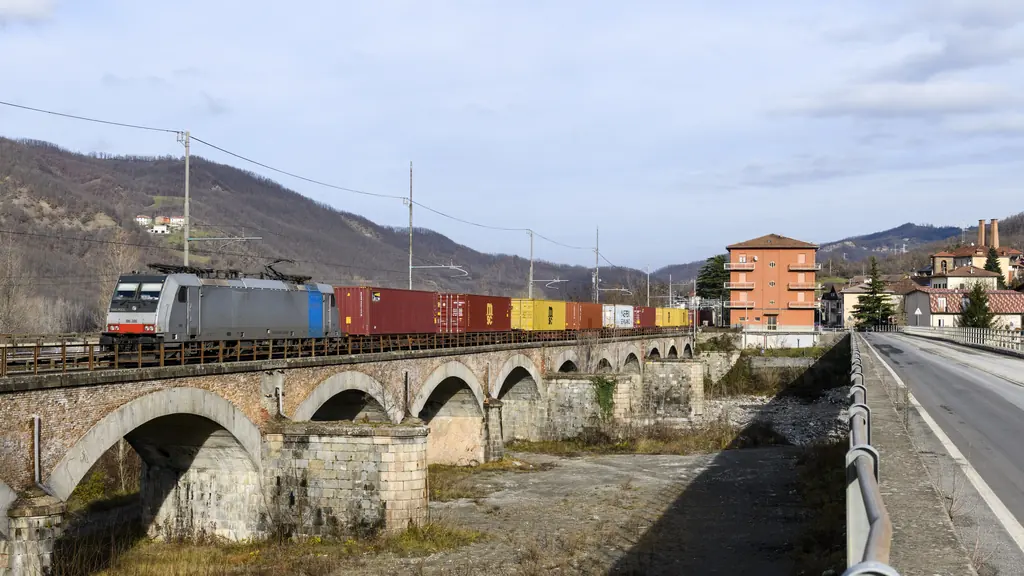 An electric locomotive hauling a container train across a stone viaduct in hill country.