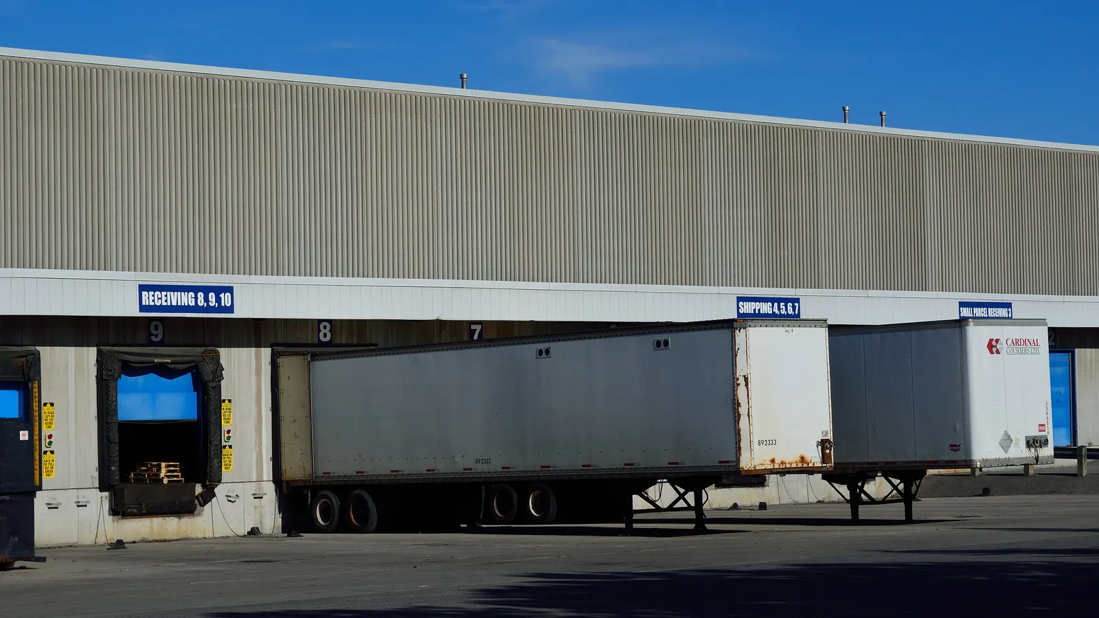 Semi-trailers backed onto numbered loading bays along a warehouse wall, with the doors signed for shipping and receiving and pallets visible inside one open bay.