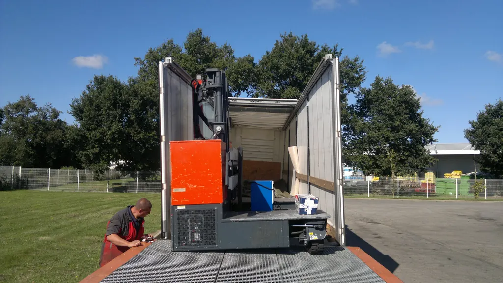 A machine on a pallet being loaded into the back of a curtainsider over a dock plate, with the rear doors open and the roof raised.