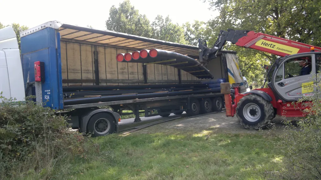 A telehandler lifting a bundle of long pipes into the open side of a curtainsider semi-trailer.