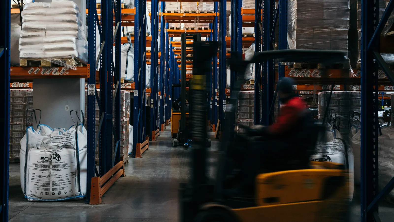 A counterbalance forklift moving down an aisle between loaded pallet racking, with bulk bags and palletised goods on the racks either side.